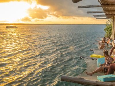 Menschen entspannen auf einer Holzterrasse mit Blick auf das ruhige Wasser bei Sonnenuntergang.