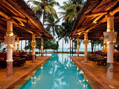 Ein tropischer Pool mit Palmen und beleuchtetem Holzrestaurant bei Sonnenuntergang.