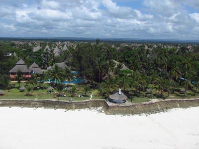Vue aérienne d'une station avec plage, palmiers, piscines et bâtiments sous un ciel nuageux.