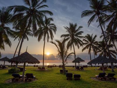 Sonnenuntergang am Strand mit Palmen und Liegestühlen unter Strohdächern.