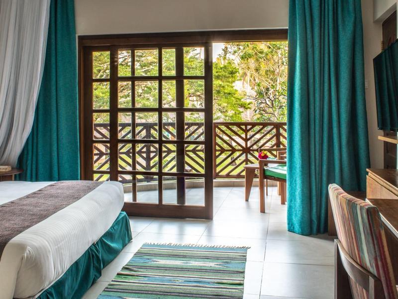 Chambre d'hôtel avec lit double, rideaux verts et balcon avec rampe en bois.