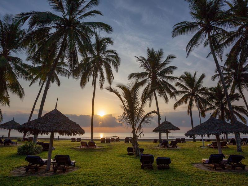 Coucher de soleil sur la plage avec des palmiers, des chaises longues et des toits de chaume.
