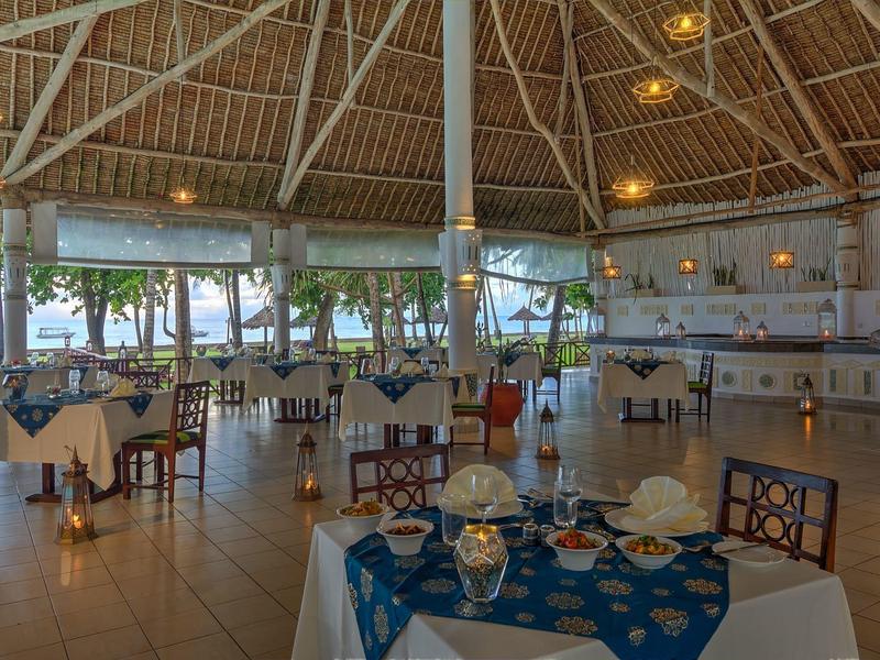 Restaurant avec vue sur la mer, tables avec nappes bleues et repas dressé sous un toit élevé.