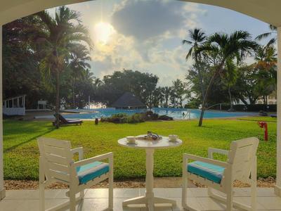 View from a covered terrace with white chairs overlooking a pool and palm trees at sunset.