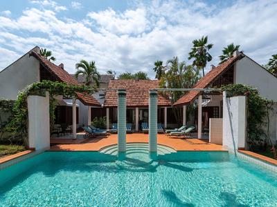 View of a pool with waterfalls in front of a tropical hotel with red roofs and palm trees.