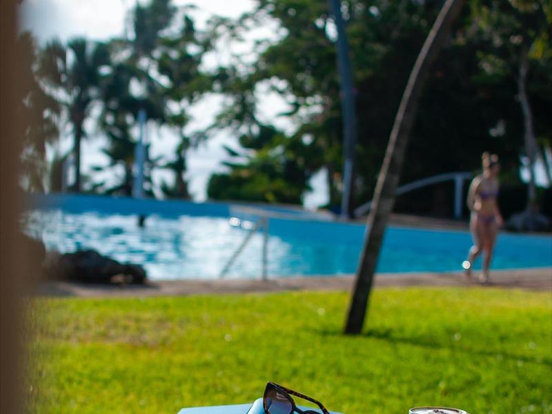 Small white table with two cups, sunglasses, and book, view of pool and palm trees.
