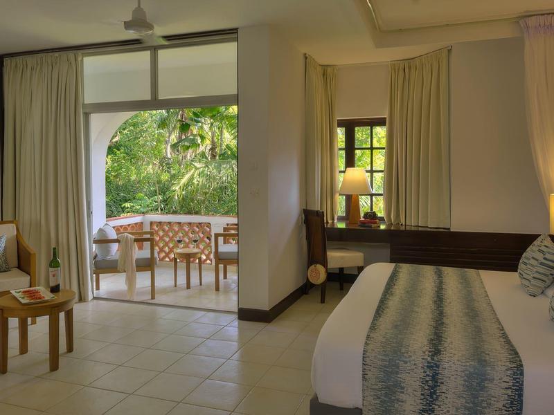 Bright hotel room with bed, chair, and view of a terrace with table and chairs.