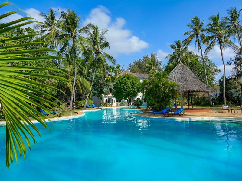 Blue pool surrounded by palm trees and tropical plants under a blue sky at a resort.