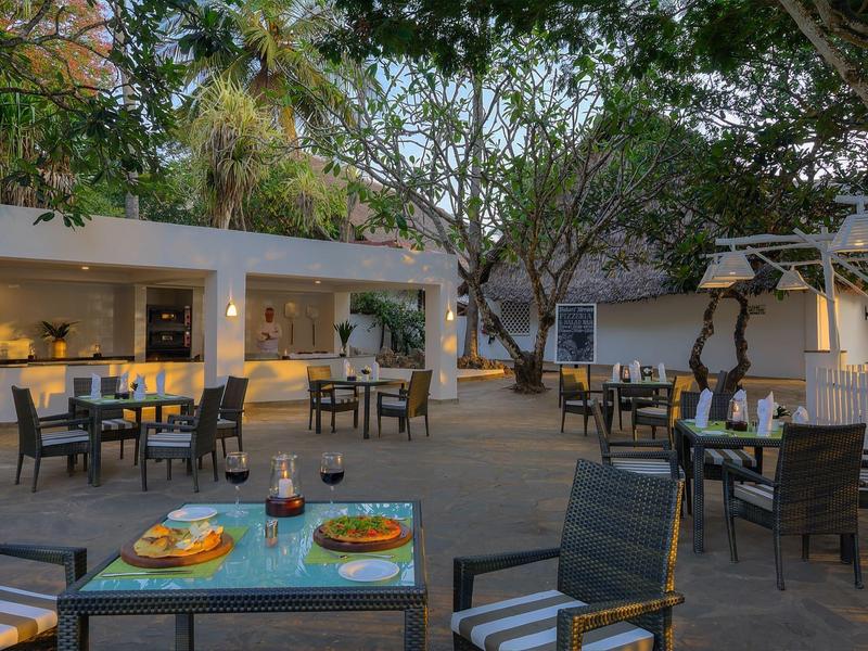 Outdoor restaurant area with tables and chairs under trees in daylight.