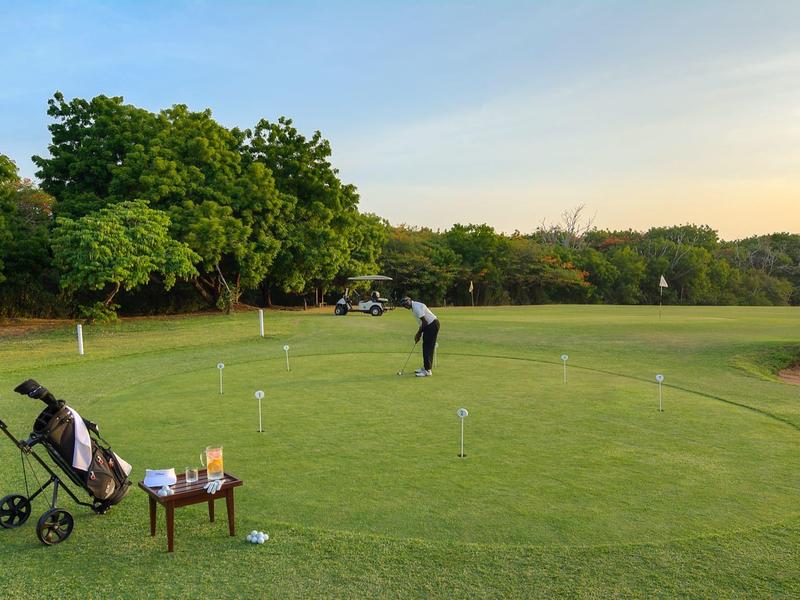 Green golf course with player, golf bag on cart, and small table with drinks.