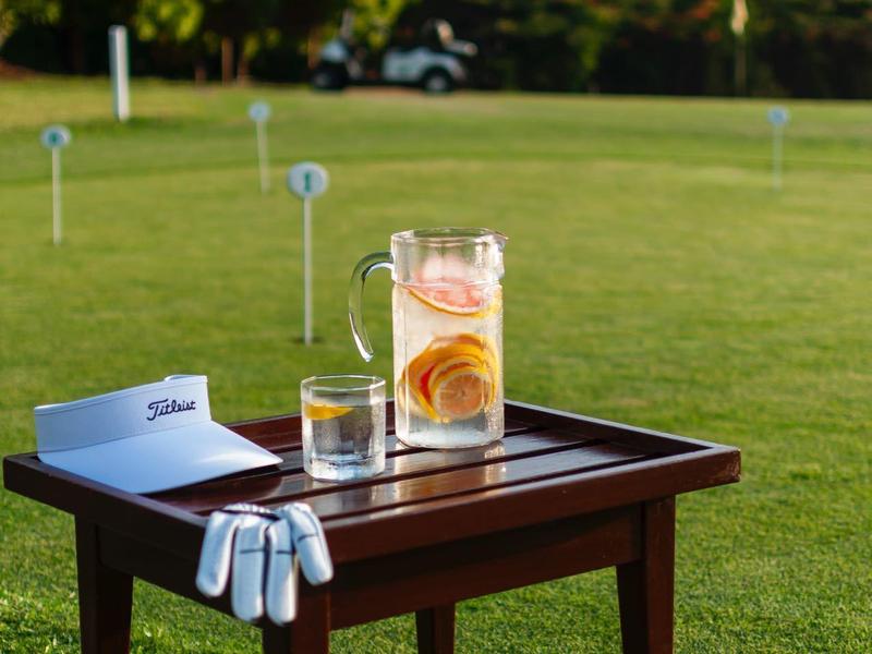 A wooden table on a green golf lawn with a drink, ball holder, and golf balls.