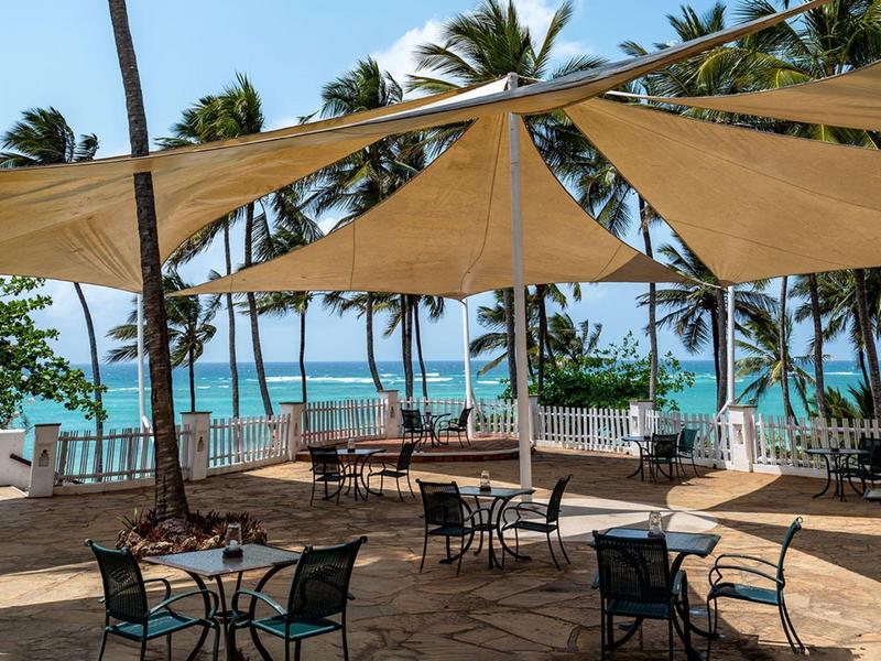 Terrace with tables and chairs under sun sails overlooking the sea and palm trees.