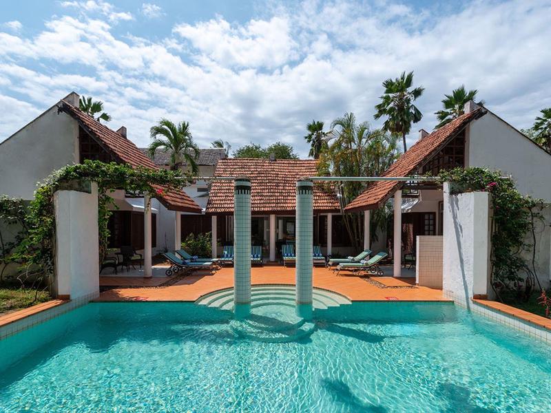 View of a pool with waterfalls in front of a tropical hotel with red roofs and palm trees.