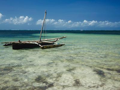 Kleines Holzboot mit Mast schwimmt im klaren, flachen Wasser unter blauem Himmel mit Wolken.