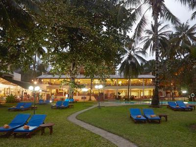 Wieczorny widok tropikalnego ogrodu hotelowego z niebieskimi leżakami pod palmami i oświetlonym budynkiem.