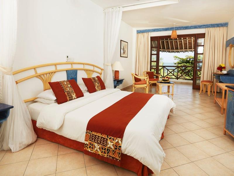 Bright hotel room with large bed, seating area, and terrace access with sea view.