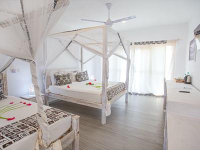 Chambre d'hôtel lumineuse avec lit double et simple, moustiquaire et balcon avec rideaux.