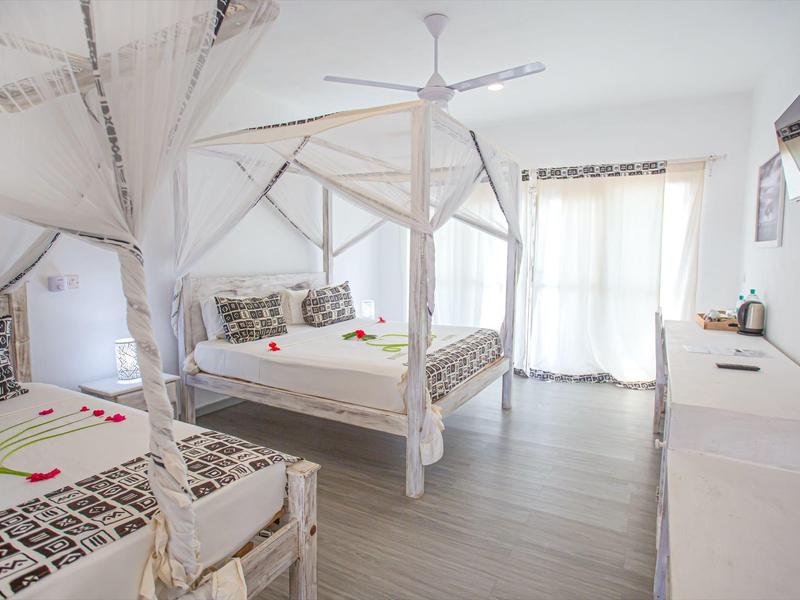 Chambre d'hôtel lumineuse avec lit double et simple, moustiquaire et balcon avec rideaux.