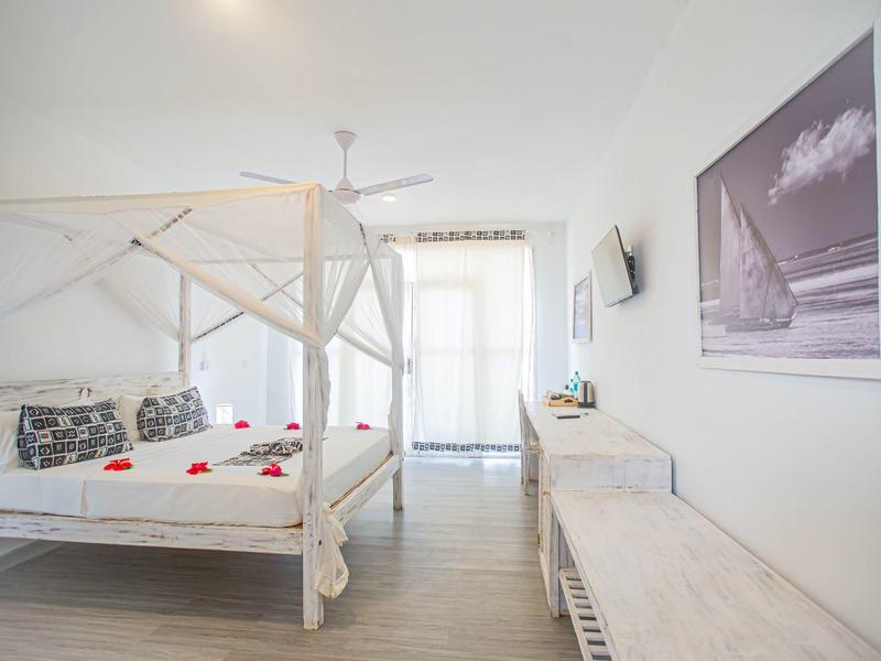 Chambre d'hôtel lumineuse avec lit à baldaquin, ventilateur et décor moderne aux tons blancs.