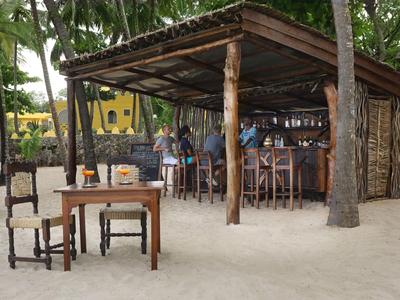 Otwarty bar na plaży z dachówką z trzciny i drewnianymi meblami na piasku, w tle drzewa i budynki.