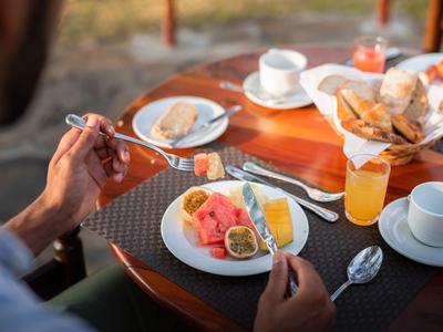 Persona con tenedor y cuchillo, desayunando frutas tropicales y jugo en mesa redonda.
