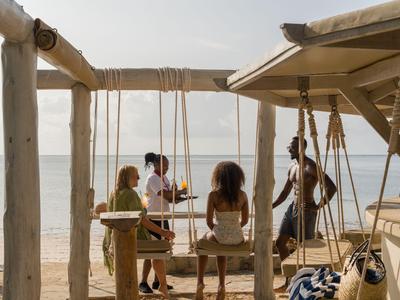 Menschen sitzen auf Schaukeln unter einem schattenspendenden Holzpavillon am Strand.