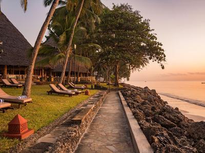 Ein Küstenweg entlang eines Resorts mit Liegestühlen, Palmen und ruhigem Meer bei Sonnenuntergang.