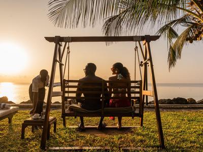 Paar sitzt auf einer Schaukel am Strand mit Sonnenuntergang und Palmen im Hintergrund.