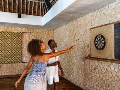 Zwei Urlauber spielen Darts in einem rustikalen Raum mit Steinhänden und Holzboden.