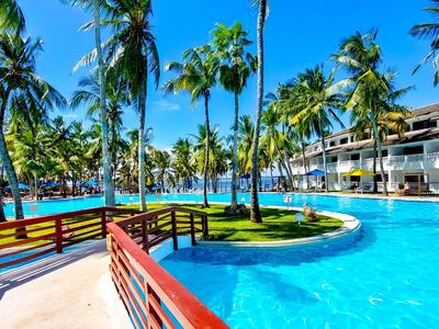 Large hotel pool with palm trees, pedestrian bridge, and modern buildings in the background under sunny weather.