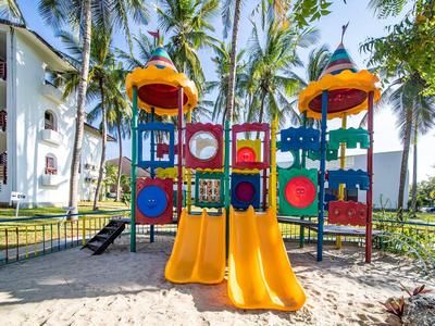 Bunter Spielplatz mit Rutschen und Klettergerüst in sandigem Bereich, umgeben von Palmen und Häusern.