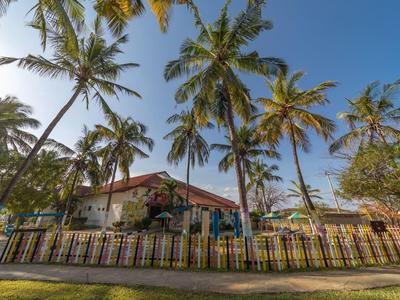 Haus mit rotem Dach hinter hohen Palmen und einem bunten Zaun bei strahlend blauem Himmel.