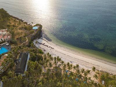 Strand mit palmenreicher Küste, weißen Sand und türkisfarbenem Meer, dahinter Hügel mit Gebäuden.