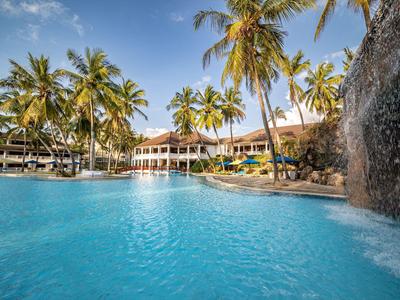 Klare blaue Poollandschaft mit Wasserfall, umgeben von Palmen und einem großen Hotelgebäude.