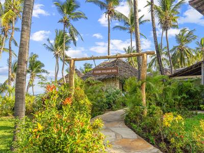 A paved path leads through tropical plants and palm trees to a beach hut under a blue sky.