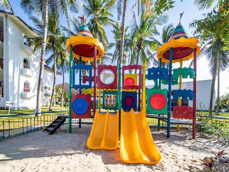 Bunter Spielplatz mit Rutschen und Klettergerüst in sandigem Bereich, umgeben von Palmen und Häusern.