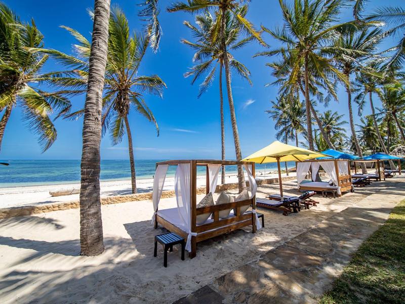 Strand mit weißen Sand, Palmen, und Holzliegen unter gelben Sonnenschirmen am blauen Meer.