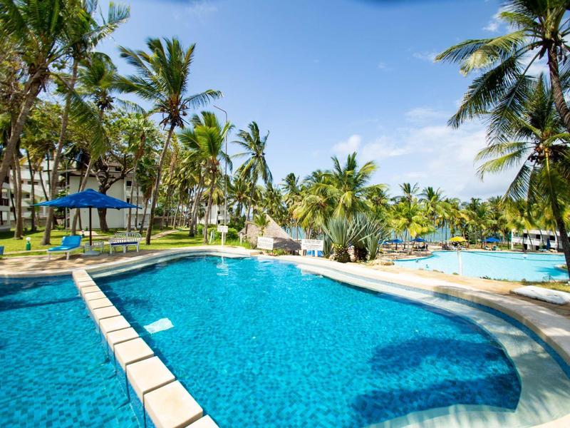 Großer blauer Pool mit Palmen und blühendem Gelände, blauem Himmel und Hotelgebäude im Hintergrund.