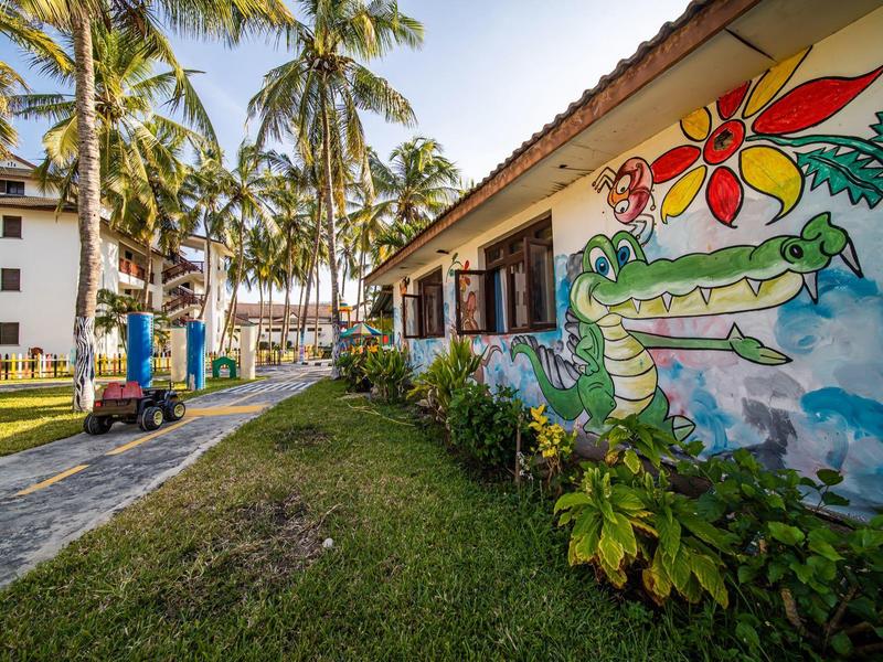 Tropischer Garten mit Palmen, buntem Krokodil-Wandgemälde und Gebäude in warmem Sonnenlicht.