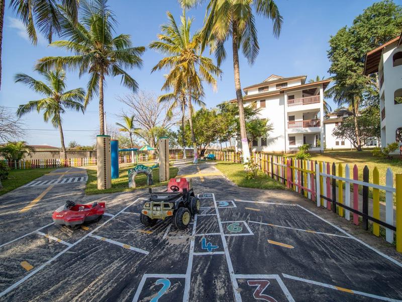 Spielplatz mit Schaukel, Rutschauto, viele Palmen und farbige Zäune bei sonnigem Himmel.