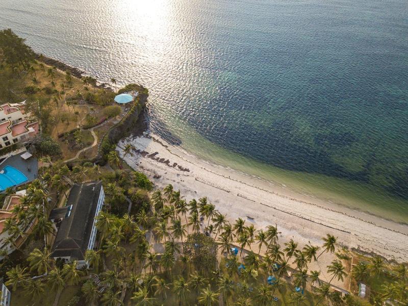 Strand mit palmenreicher Küste, weißen Sand und türkisfarbenem Meer, dahinter Hügel mit Gebäuden.