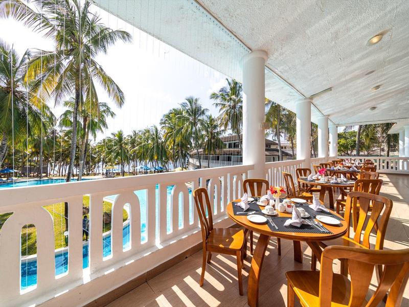 Balkon mit Holztischen und Stühlen, Blick auf Palmen und Pool unter bewölktem Himmel.