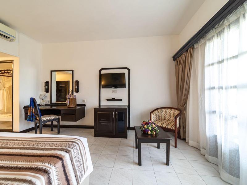 Helles Hotelzimmer mit Doppelbett, Sessel, Tisch, Fernseher und großem Fenster mit Vorhängen.