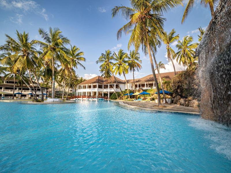 Klare blaue Poollandschaft mit Wasserfall, umgeben von Palmen und einem großen Hotelgebäude.