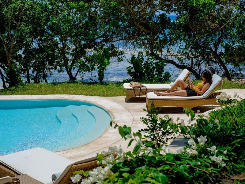 Personnes se détendant sur des chaises longues près de la piscine avec arbres et mer en arrière-plan.