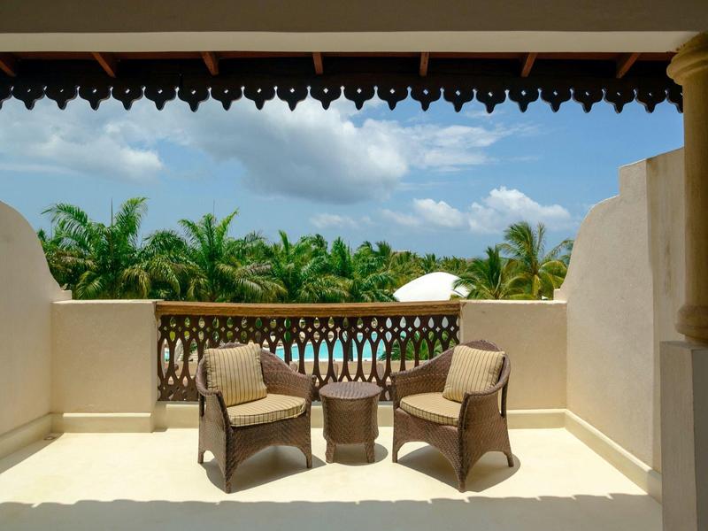 Balcon avec deux fauteuils et une table, vue sur des palmiers et un ciel bleu.