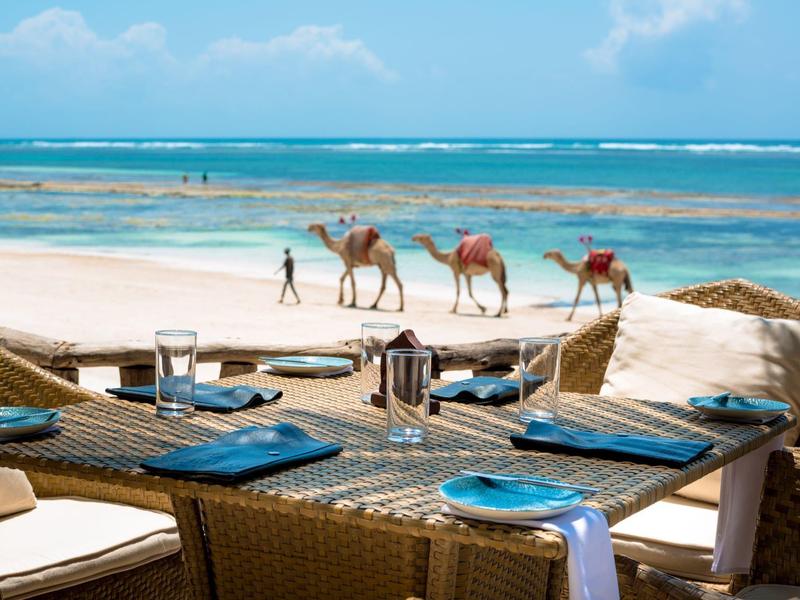 Espace repas sur la plage avec une caravane de chameaux en arrière-plan par ciel clair.