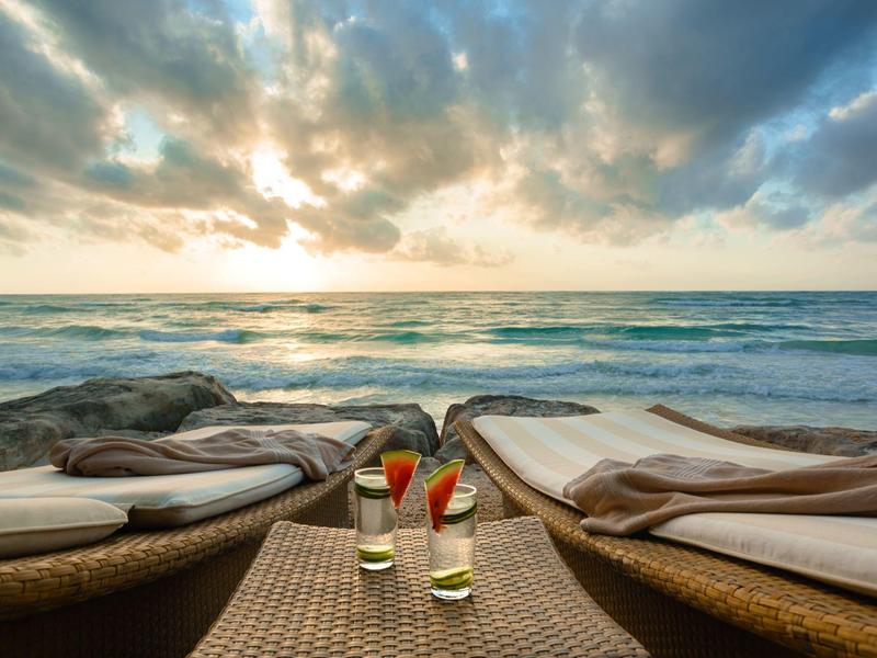 Deux chaises longues avec coussins face à la mer au coucher du soleil et deux boissons sur une table.