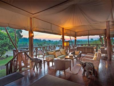Offene Veranda mit Holzgeländer, Sesseln und Tischen mit Blick auf grüne Landschaft bei Sonnenuntergang.