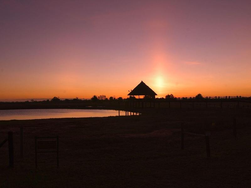 Atardecer con cielo púrpura-naranja sobre un lago con la silueta de una cabaña en la orilla.